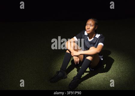 African American female athlete sitting on turf under spotlight, wearing soccer jersey and cleats Stock Photo