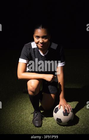 African American woman kneeling on soccer pitch wearing black kit holding ball under spotlight. Athlete, footballer, night, dramatic, jersey, cleats, Stock Photo