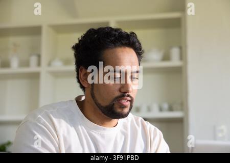 African man sitting at kitchen counter wearing light shirt, working on laptop with open shelving Stock Photo