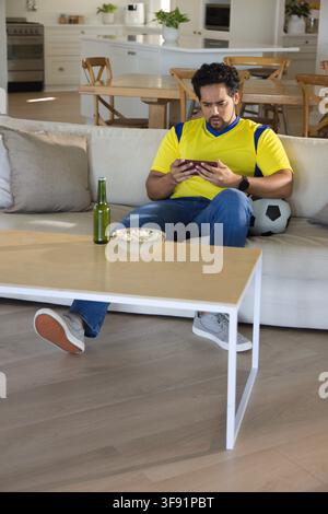 Mid-adult African man sitting on sofa in home wearing yellow-jersey holding smartphone, soccer ball Stock Photo