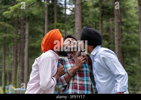 Waist-up shot of three male friends, including two Sikh men, standing closely together and having fun in a pine forest in Himachal Pradesh. Stock Photo