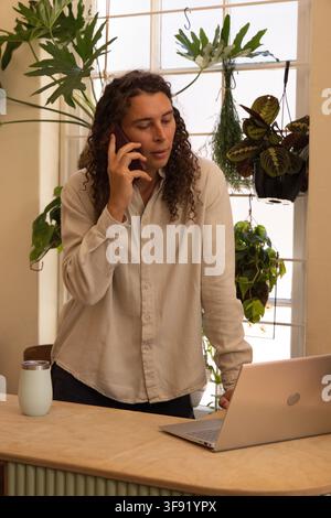 Non-binary person standing behind light wood countertop at home, holding phone, leaning by laptop. Remote, workspace, plants, daylight, greenery, prod Stock Photo