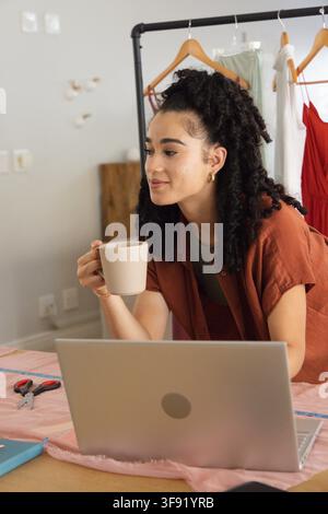 Woman in rust shirt, leaning over studio table with pink fabric, holding mug and checking laptop. Sewing, seamstress, designer, textiles, sketchbook, Stock Photo
