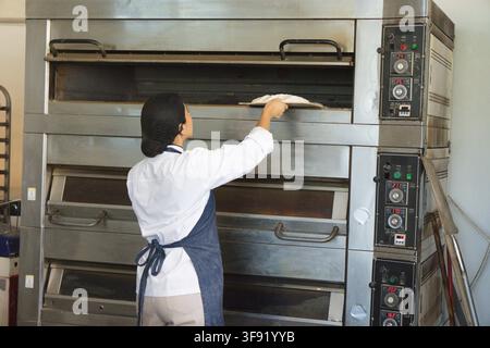 Female baker wearing chef jacket and apron placing dough using peel at commercial bakery oven. Kitchen, industrial, stainless, production, breadmaking Stock Photo