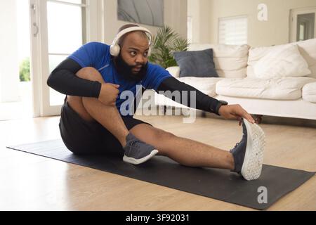 African American man doing hamstring stretch on mat at home wearing blue shirt, headphones, tracker Stock Photo