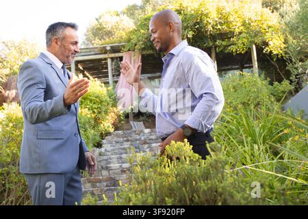 Diverse men greeting on stone steps in garden, one wearing suit, other wearing shirt and watch. Outdoor, patio, pergola, greenery, sunlight, blue, tie Stock Photo