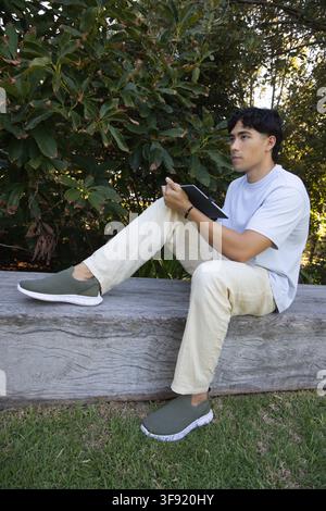 Asian male sitting on weathered wooden bench in park holding tablet and using stylus Stock Photo