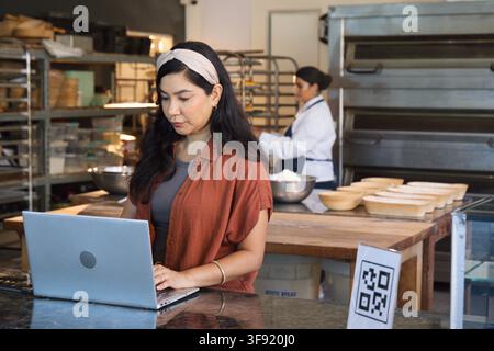 Diverse female coworkers at counter using laptop, baker in chef jacket handling mixing bowls. Bakery, workspace, teamwork, baking, workflow, productiv Stock Photo