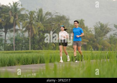 Asian Indonesian couple running on a narrow paved path through vibrant rice fields with tropical trees and mountain scenery in Nanggulan, Yogyakarta. Stock Photo
