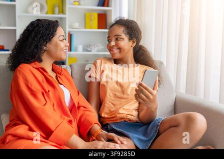 Black mother and daughter smiling at each other while looking at phone Stock Photo