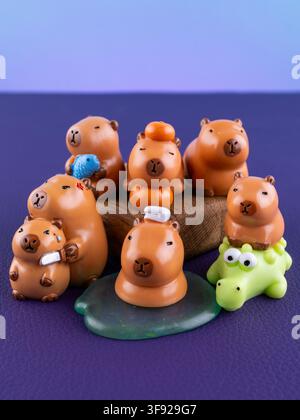 Close-up of cute miniature capybara figurines in different poses with oranges and a tiny crocodile on a vibrant purple background. Adorable decorative Stock Photo