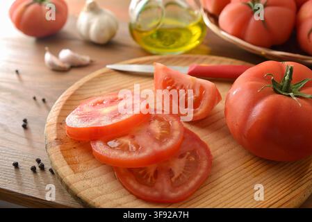 Fresh tomatoes are sliced on a wooden cutting board with garlic, olive oil, and a knife, creating a warm rustic kitchen scene. Stock Photo