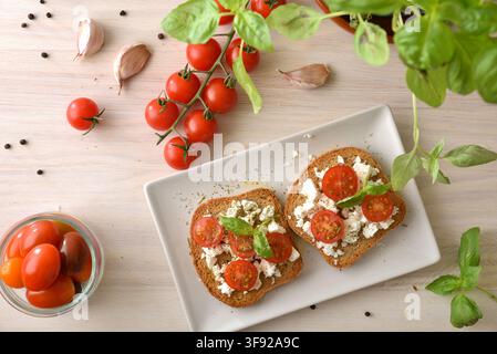 Two feta-topped toasts with sliced tomatoes and basil are arranged on a white plate, surrounded by garlic, peppercorns and fresh ingredients on a wood Stock Photo