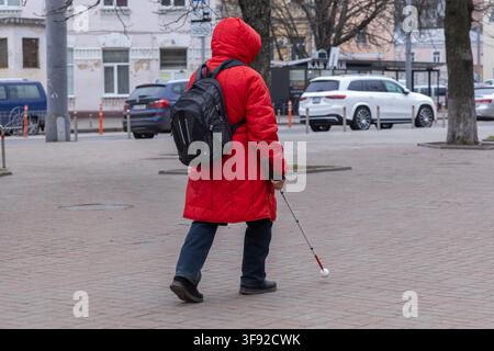 Kyiv, Ukraine - March 22, 2026: The person wearing a red hooded coat navigates a paved city area using a long cane for guidance, highlighting the impo Stock Photo