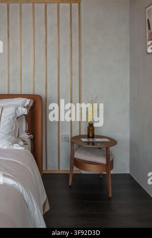 Cozy minimalist bedroom corner featuring a round wooden nightstand decorated with yellow flowers in a glass bottle, books, and folded blanket. Stock Photo