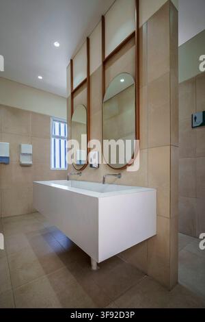 Contemporary public washroom interior featuring a large floating white double sink trough with dual chrome faucets. Above the vanity counter. Stock Photo
