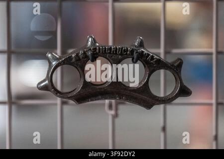 VIENNA, AUSTRIA, 13 August 2026: A dark metal brass knuckles weapon hangs against a wire grid with a blurred background. Stock Photo