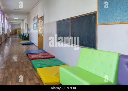 A long brightly lit school hallway features vibrant soft mats on wooden floorboards and tufted chairs for children, capturing an inviting atmosphere o Stock Photo