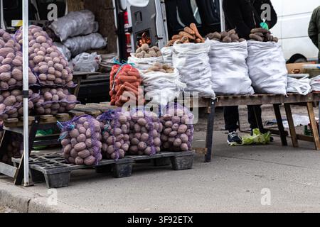Assorted potatoes, carrots, and beets sold in large quantities at a sidewalk farm stall, emphasizing seasonal food availability and a rustic community Stock Photo