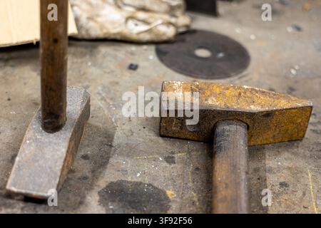 Two well used hammers with wooden handles and rusty metal heads lie on a dirty workshop table, surrounded by various industrial tools and debris refle Stock Photo