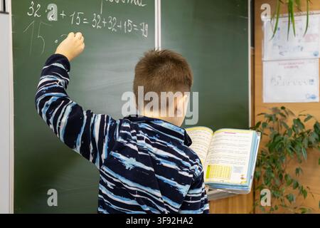 Kyiv, Ukraine - March 20, 2026: A focused schoolboy solves difficult math problems on a classroom blackboard using a reference book to illustrate his Stock Photo