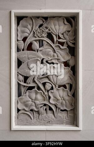 Detailed stone relief wall sculpture depicting a graceful heron bird standing amidst lotus flowers, leaves, and seed pods. Carved with intricate Asian Stock Photo