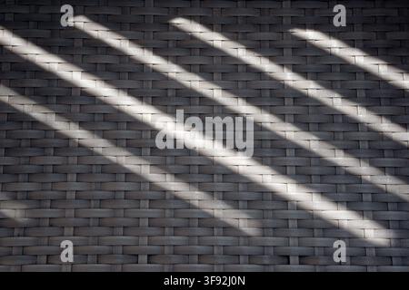 Close-up texture background of a grey woven synthetic rattan surface illuminated by bright natural sunlight creating parallel diagonal shadows. Stock Photo