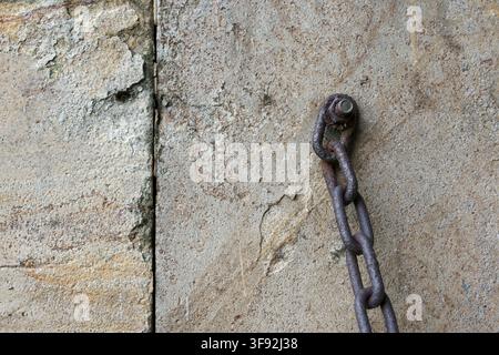 Close-up shot of a heavy, rusted metal iron chain hanging from a bolt embedded in an aged concrete wall. The weathered grey cement texture and oxidize Stock Photo