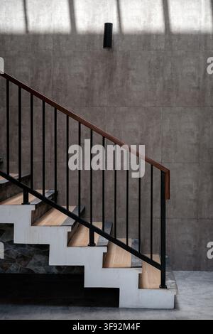 Interior shot of a stylish modern staircase featuring light wood steps, black metal handrail, white zig-zag structure, and textured grey wall lighting Stock Photo