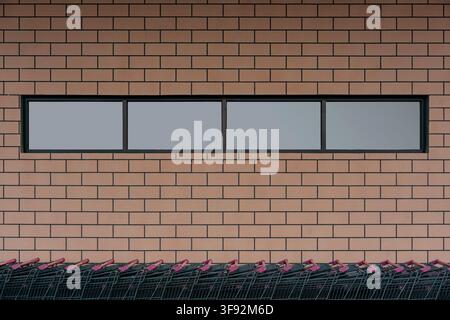 A straight front view of dark metal shopping carts with red handles lined up below a horizontal window on a brown brick building facade. Stock Photo