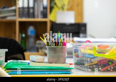 Assorted artistic tools held in a storage cup next to a bin filled with pencils and scissors on a cluttered desktop, depicting a busy environment for Stock Photo