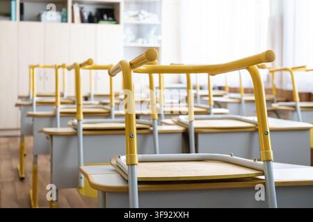 Rows of school furniture organized in a sunlight lit classroom with chairs placed upon tables, capturing a clean and prepared environment for students Stock Photo