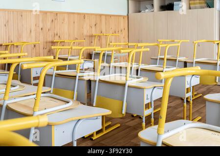 Rows of wooden student desks with yellow metal frames are arranged in a tidy classroom setting where chairs are inverted to signify a school holiday p Stock Photo