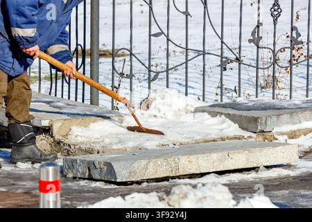 A worker is using a tool to clear snow from the sidewalk in a cold weather scene. The worker is dressed in a blue jacket and brown pants. There are la Stock Photo