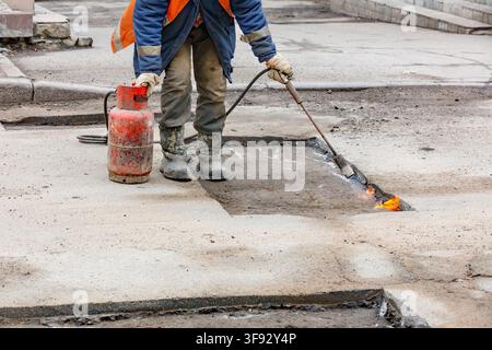 A worker stands on an urban street, using a torch to repair a section of the road. Flames are visible as the worker applies heat to the surface. Stock Photo