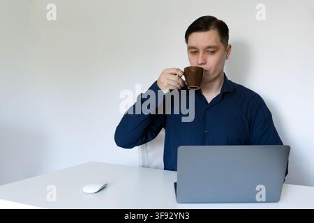 A young professional man in a dark blue shirt drinking coffee from a brown cup while looking at a grey laptop at a clean white office desk. Stock Photo