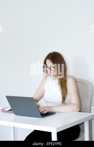 A young woman wearing glasses and a white top, sitting at a clean minimalist white desk and focused on typing on a grey laptop. Stock Photo