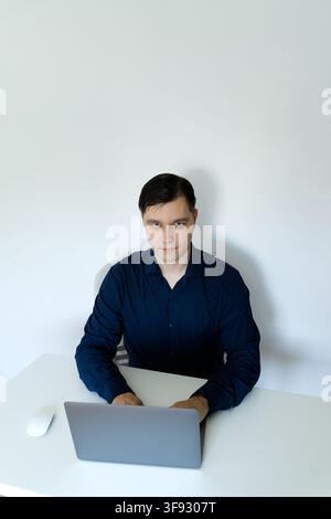 A young professional man in a dark blue shirt sitting at a clean minimalist white desk, looking directly at the camera while working on a grey laptop. Stock Photo