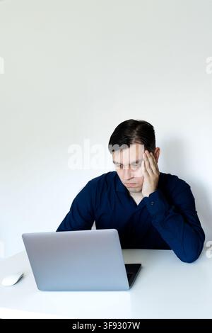 A young professional man in a dark blue shirt looking tired, stressed, and thoughtful while sitting at a clean white desk with a laptop Stock Photo