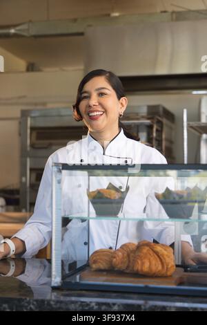 Mid-adult female chef wearing chef coat working behind glass case showing croissants at bakery Stock Photo