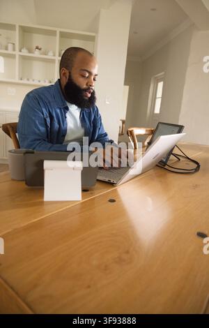 Bearded man wearing denim overshirt sitting at wooden dining table typing on laptop with tablet Stock Photo