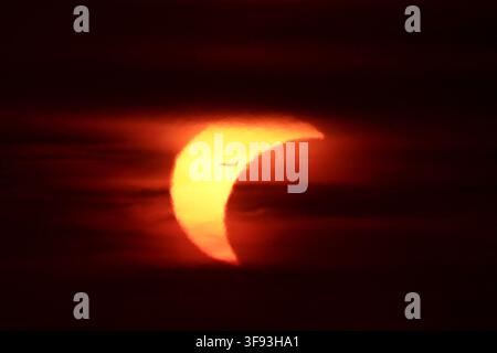 ROME, ITALY - AUGUST 12, 2026: Silhouette of a commercial airplane flying across the crescent sun during a partial solar eclipse at sunset, framed by dark atmospheric clouds. Stock Photo