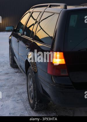 Black station wagon parked on snowy surface with illuminated turn signal. Warm golden light from the setting sun reflects off the car's body and windo Stock Photo