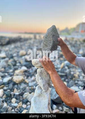 Individual carefully places a stone atop a sculpture made of balanced rocks on a beach, with the ocean and buildings in the distance Stock Photo
