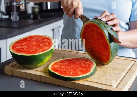 A large knife cuts through a halved watermelon standing on a wooden cutting board, with another half and a round slice nearby. Stock Photo