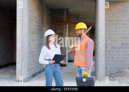 Architect and foreman wearing protective workwear discussing building project using spirit level and clipboard Stock Photo
