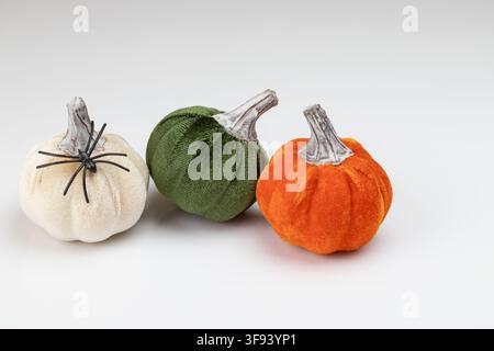 A horizontal Halloween-themed still life featuring three velvet pumpkins in white, green, and orange. A small black decorative spider is placed on top Stock Photo