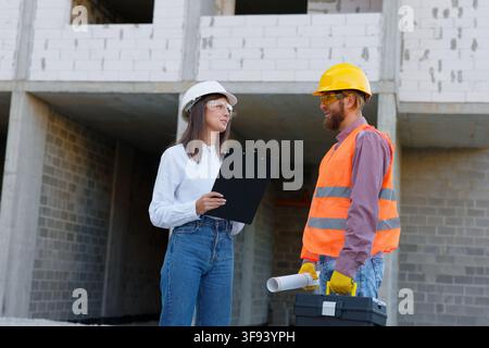 Female architect holding clipboard and male construction worker carrying blueprints and toolbox are inspecting building progress Stock Photo