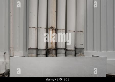 A front view of several vertical white industrial drain pipes with weathered rust marks mounted against a corrugated metal wall above a concrete base. Stock Photo