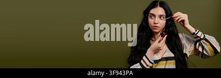 Elegant young woman poses gracefully, applying beauty serum on face against an olive backdrop, banner Stock Photo